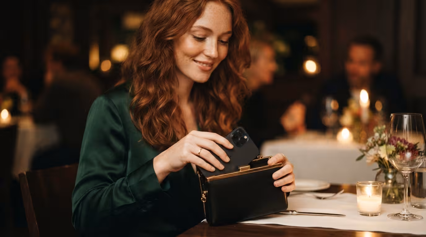 A young woman gracefully slipping her phone into her clutch bag as she settles at a restaurant table